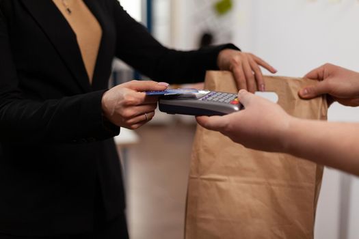 Close up of business woman holding credit card of pos, using contactless technology, pay for takeaway food from courier. Holding box with meal lunch in paper bag.