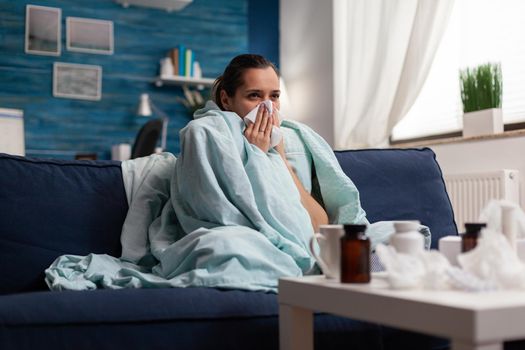 Woman with sickness wrapped in blanket at home ill feeling unwell. Young caucasian adult suffering from temperature fever disease illness flu, taking medical treatment sitting on couch