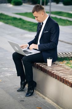 Distance working. Isolated man in a suit. Handsome young businessman sitting outdoors working with his laptop. Crisis 2020. Coronavirus outbreak.