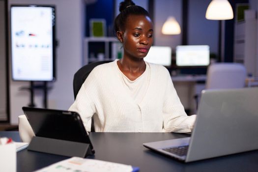 Workaholic african manager trying to finish deadline checking charts on tablet pc and laptop. Busy multitasking employee analysing financial statistics overworking writing, searching.