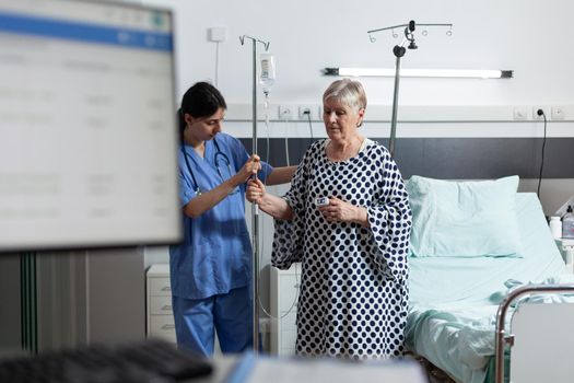 Sick senior woman carrying iv drip bag with help from medical nurse in hospital room, receiving a medication through an intravenous line. Blood oxygen saturation measured with oxymeter.