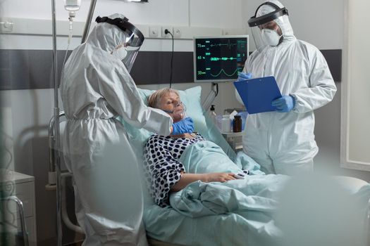Medical staff in ppe suit helping patient breath with oxygen mask, because of pulmonary failure, dressed in coverall during coronavirus outbreak. Getting intravenous medicine from iv bag.