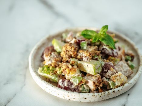 Waldorf salad with copy space. American fruit and nut salad with apples, celery, grapes, chicken meat, dressed mayonnaise. Craft plate with ready-to-eat waldorf salad on white marble background