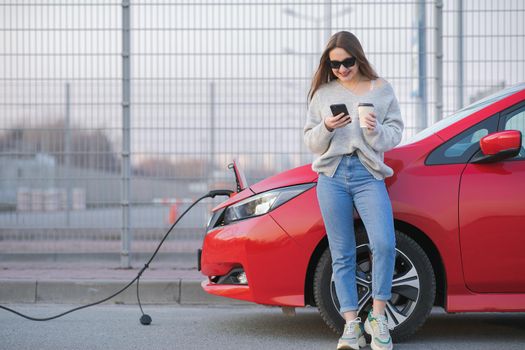 Electric car charging in street. Ecological Car Connected and Charging Batteries. Girl Use Coffee Drink While Using SmartPhone and Waiting Power Supply Connect to Electric Vehicles for Charging.