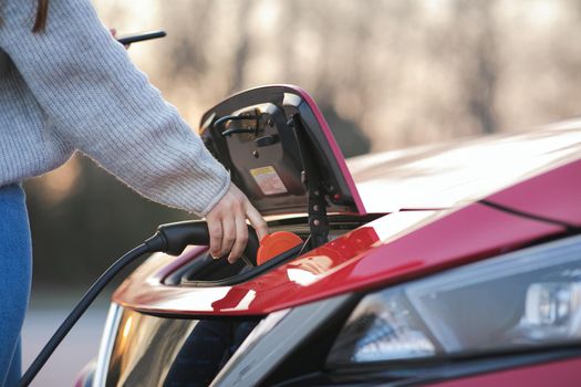 Woman is plugging electric vehicle for charging car battery at parking. Close up. Plugged charging cable electric vehicle, EV parking, power charger cable, charging port station, sustainable future