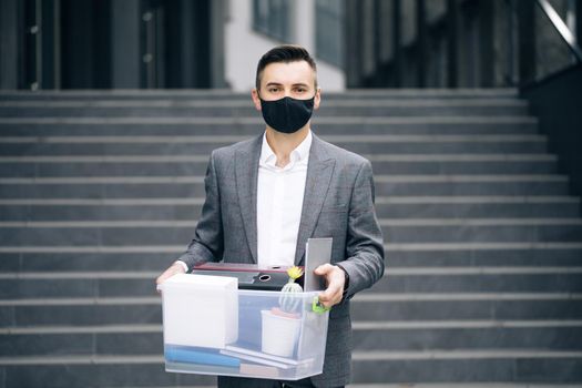 Fired sad young businessman in medical mask carries box with personal belongings leaving the office. Unemployment. Lack of jobs. Financial crisis