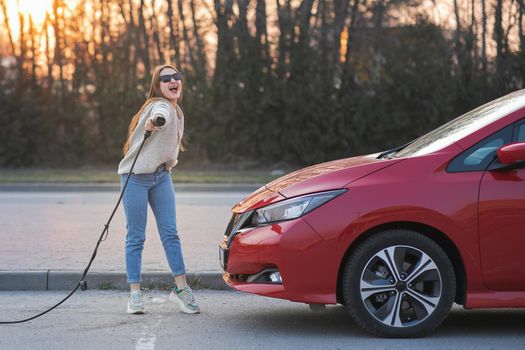 Happy young girl holding power cable supply in hand standing near electric automobile and looking at camera