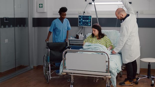 Therapist doctor monitoring heart pulse during medical recovery in hospital ward. Black woman nurse putting sick patient in wheelchair for physiotherapy rehabilitation recovering for leg accident