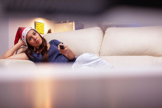 Woman adult resting on couch watching xmas movie on television during christmastime in x-mas decorated kitchen. Person wearing red santa hat enjoying winter season celebrating christmas holiday