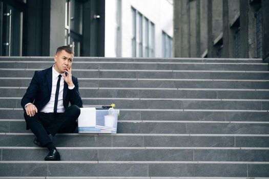 Sad businessman sitting on stairs outdoor with box of stuff as lost business. Fired man. Unemployment rate growing due pandemic. Male office worker in despair lost job.