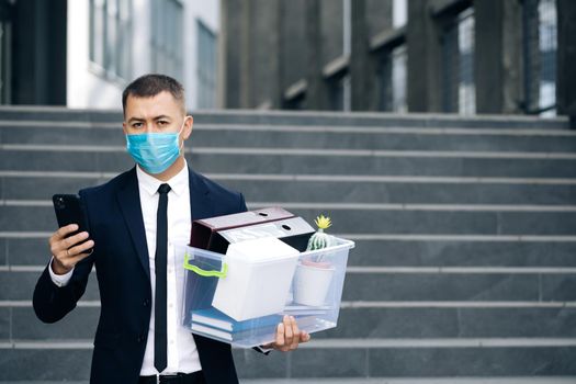 Businessman in medical mask with box of personal stuff walking the street use phone. Business style suit. Coronavirus outdoors social distancing. Finance and industry. Fired man lost job.
