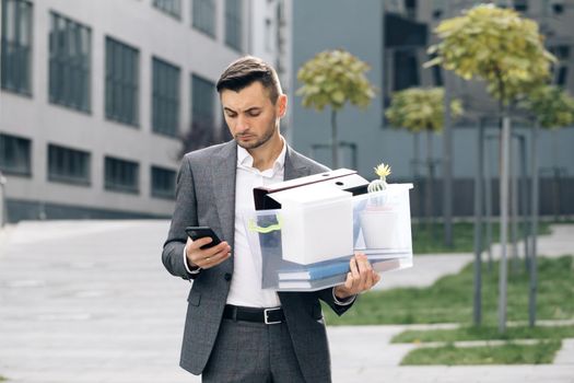 Sad young businessman uses phone texting scrolling tapping near office building. Male office worker in despair lost job. Look serious technology communication.