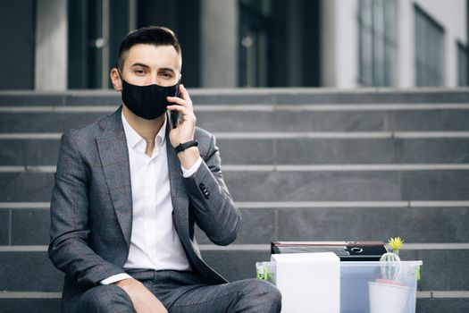 Fired man in medical mask talking on phone with stuff in box. Cellphone conversation. Speaking on telephone. Handsome jobless office worker entrepreneur in formal suit sitting on stairs outdoors.