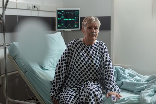 Senior woman patient sitting on the edge of bed in hospital bed looking pensive, breathing inhale exhale with help from oxygen mask, receiving medication through an intravenous line