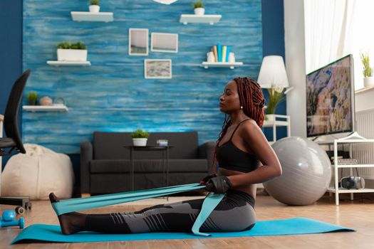 Black woman doing pilates workout using elastic strap sitting on yoga mat, pulling training arms and shoulders in home living room. Athletic fit exercising body using resistance band.