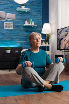 Senior woman in sportwear warming up abdominals muscles practicing gym body exercise using workout dumbbells. Pensioner sitting on yoga mat in lotus position during wellness routine in living room