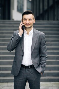 European businessman talking on mobile phone near office building background. Man is in dialogue, smiling, holding coffee cup on summer day.