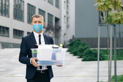 Young sad businessman in protective face mask walking down the stairs outdoor with box of stuff as leaving business center. Unemployment rate growing due pandemic. Male office worker lost her job.