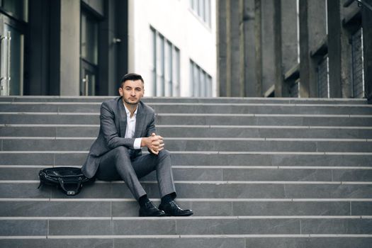 Feeling bad and annoyed. Sad young businessman sitting on the stairs near office building. Tired putting hands to the head. Male office worker in despair lost job.