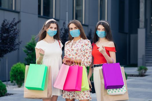 Group of young women wearing masks to protect coronavirus pandemic with colored bags in hands. Consumerism, purchases, shopping, lifestyle concept.