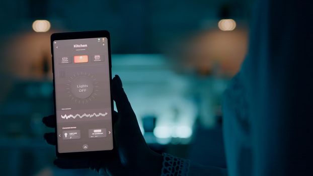 Woman holding smartphone with voice activation smart light app to turn on the lights in house. Person using future technology and smart software switching on bulbs wireless before working at laptop