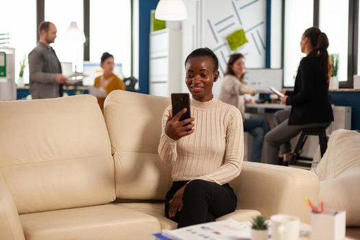 African manager woman discussing with remote colleagues on video call holding smartphone using headphones sitting on couch in business modern office. Diverse coworkers planning new financial project