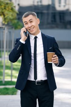 Man is in dialogue, smiling, holding coffee cup on summer day. European businessman talking on mobile phone walking near office building background.