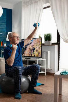 Retirement senior man sitting on aerobics swiss ball raising hand streching arm muscle doing fitness exercise. Pensioner exercising body muscles using dumbbells training muscular resistance