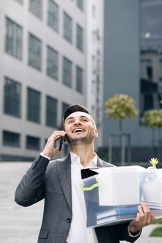 Happy fired man talking on phone with stuff in box. Speaking on telephone. Cellphone conversation. An employee between office buildings with a box and documents with a desk flower.