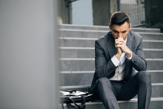 Upset young businessman in frustration outside office department. Fired unhappy caucasian bearded man losing job crying and sitting on stairs. Unemployment. Crisis.