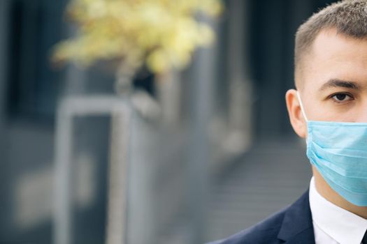 Half face of upset caucasian young handsome man in medical mask looking straight to camera while standing outdoors near modern office building. Man in protection mask look at camera.