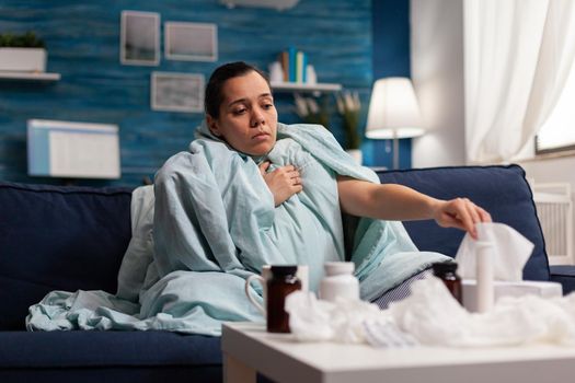 Woman with flu symptoms sitiing at home ill on couch suffering. Young caucasian person using tissues medicine treatment against fever temperature infection cold disease illness