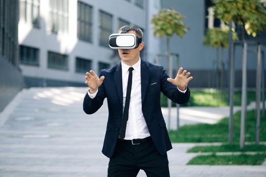 Happy businessman playing a game on imaginary screen using augmented reality goggles standing on the background of modern urban building. New technology offers new 3D dimensions