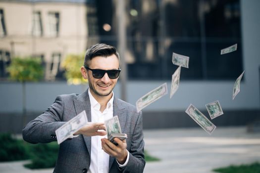 Successful Businessman in a Suit Wear Sunglasses Throwing Money in the Camera Standing in the Street near Office Building. Money rain, falling dollars. Successful business concept.
