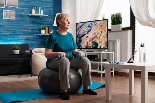 Focused senior woman doing arm exercising using workout dumbbells sitting on swiss ball in living room. Pensioner in sportswear looking at online aerobics training on tablet exercising body resistance