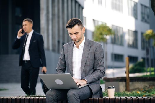Businessman typing on laptop computer outdoor. Man in suit working with laptop while sitting on bench. Remote work concept. Distance working. Isolated man on a suit.
