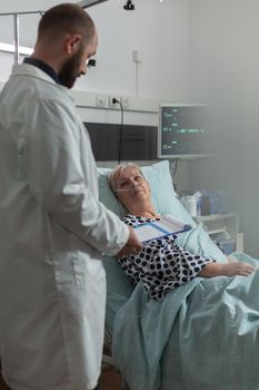 Doctor reassuring senior woman full recovery during discussion consultation in hospital room. Writing note on clipboard. Breathing with help from oxygen mask.