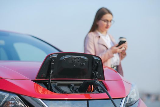 Woman stands with phone near her electric car and waits when vehicle will charged. Charging of electric car.