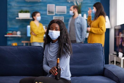 African woman keeping social distancing wearing face mask while meeting with friends to prevent spread of coronavirus holding beer bottle looking at camera sitting on couch. Conceptual image.