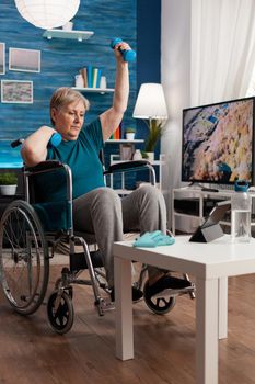 Invalid senior woman holding workout dumbbells raising arm while exercising body muscle persistence doing cardio exercise. Disabled pensioner trying recovery cardio watching online gym video on tablet