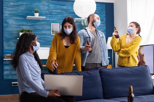 Multiracial group of friends chatting using laptop keeping social distancing and wearing face mask to prevent covid spread and infection in apartment living room. Conceptual image
