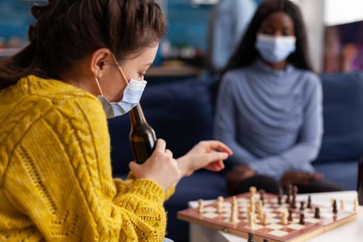 Happy woman holding beer bottle playing chess with african friend in living room keeping social distancing due to covid19 pandemic wearing face mask to prevent infection. Board games, competing, tactics, asctivity.