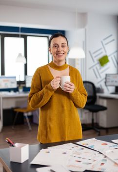 Happy confident cheerful woman looking at camera in start up company office holding cup of coffee. Executive entrepreneur, manager leader standing working on projects.