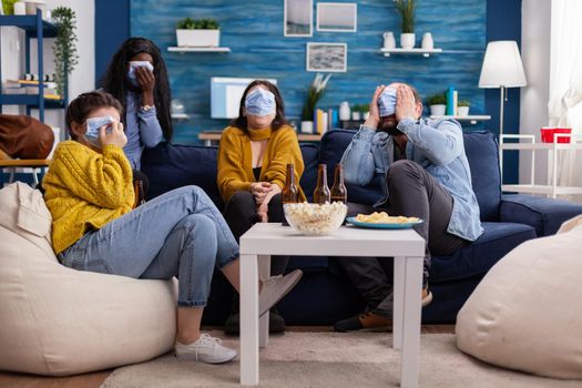 Multiethnic friends socialising looking at terrifying movie on tv sitting on sofa in apartment living room wearing face mask during coronavirus outbreak respecting social distancing.