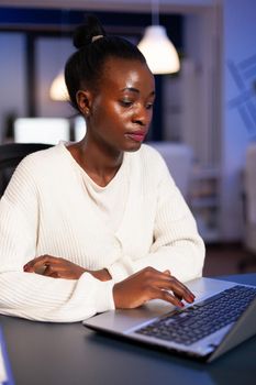 Stressed african manager woman working with financial graphs checking statistics, typing on laptop, reading raports late at night in start-up office doing overtime to respect deadline of project.