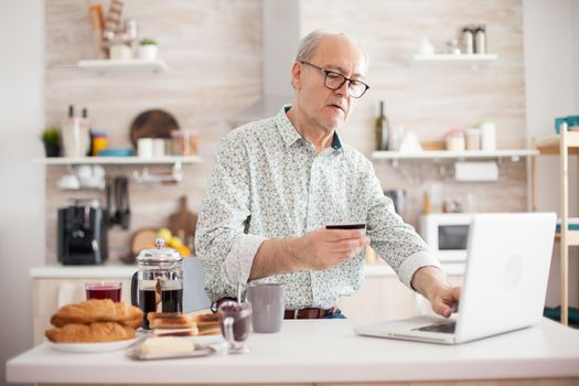 Senior man shopping online from the confort of his house. Pensioner paying online using credit card and application from laptop during breakfast in kitchen. Retired elderly person using internet payment home bank buying with modern technology