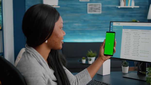 Student with dark skin holding phone with mock up green screen chroma key looking at isolated display sitting at desk table in living room. Woman texting message working at social network app