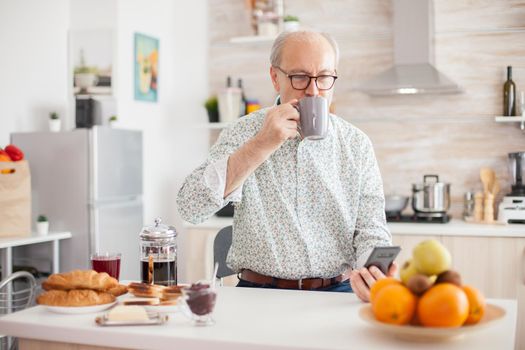 Senior man drinking coffee during breakfast in ktichen and using smartphone.