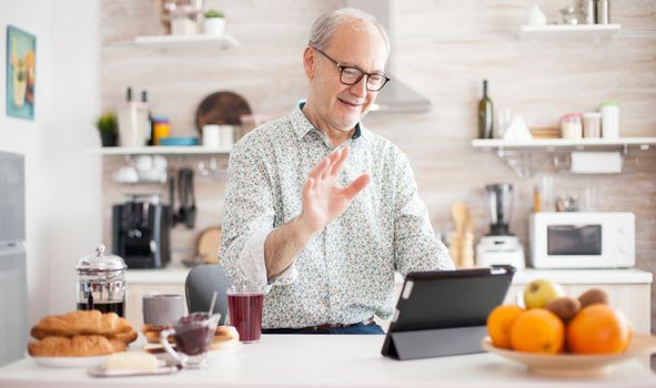 Authentic senior man on online video call in the morning in cozy kitchen. Elderly person using internet online chat technology video webcam making a video call connection camera communication conference call