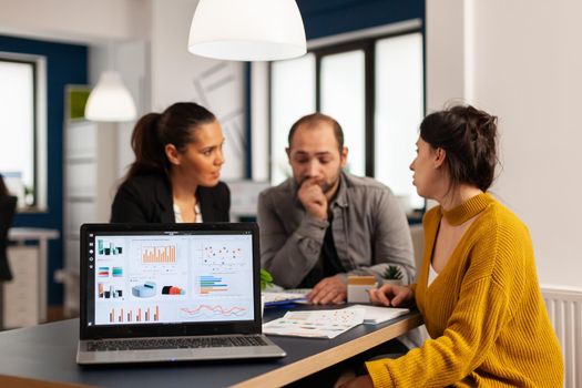 Laptop screen ready for financial project presentation placed on desk while business people work in background. Diverse employees gathered in co-working, working process in busy company, teamwork help